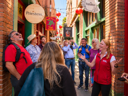 A group of tourists listening to a guide who wears a red and white branded vest while standing in Fan Tan Alley, lively street lined with brick buildings and colourful shop signs.