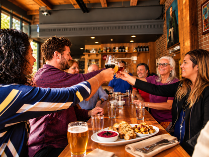 A group of friends and family sharing a joyful moment at a historical downtown Victoria Pub, clinking glasses of a local cocktail called a Shaft with plates of food on the table in a warmly lit restaurant setting.